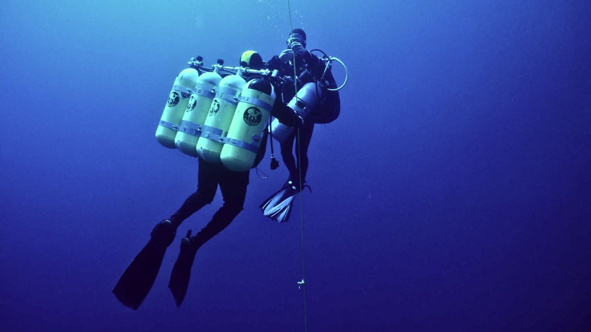 Technical diver in specialized equipment with multiple tanks attempting extreme depth record dive in dark deep blue water showing world diving record achievement